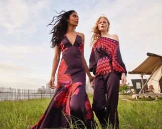 Two female models in purple printed outfits standing outdoors on grass near a waterfront skyline by photographer Juan Algarin.