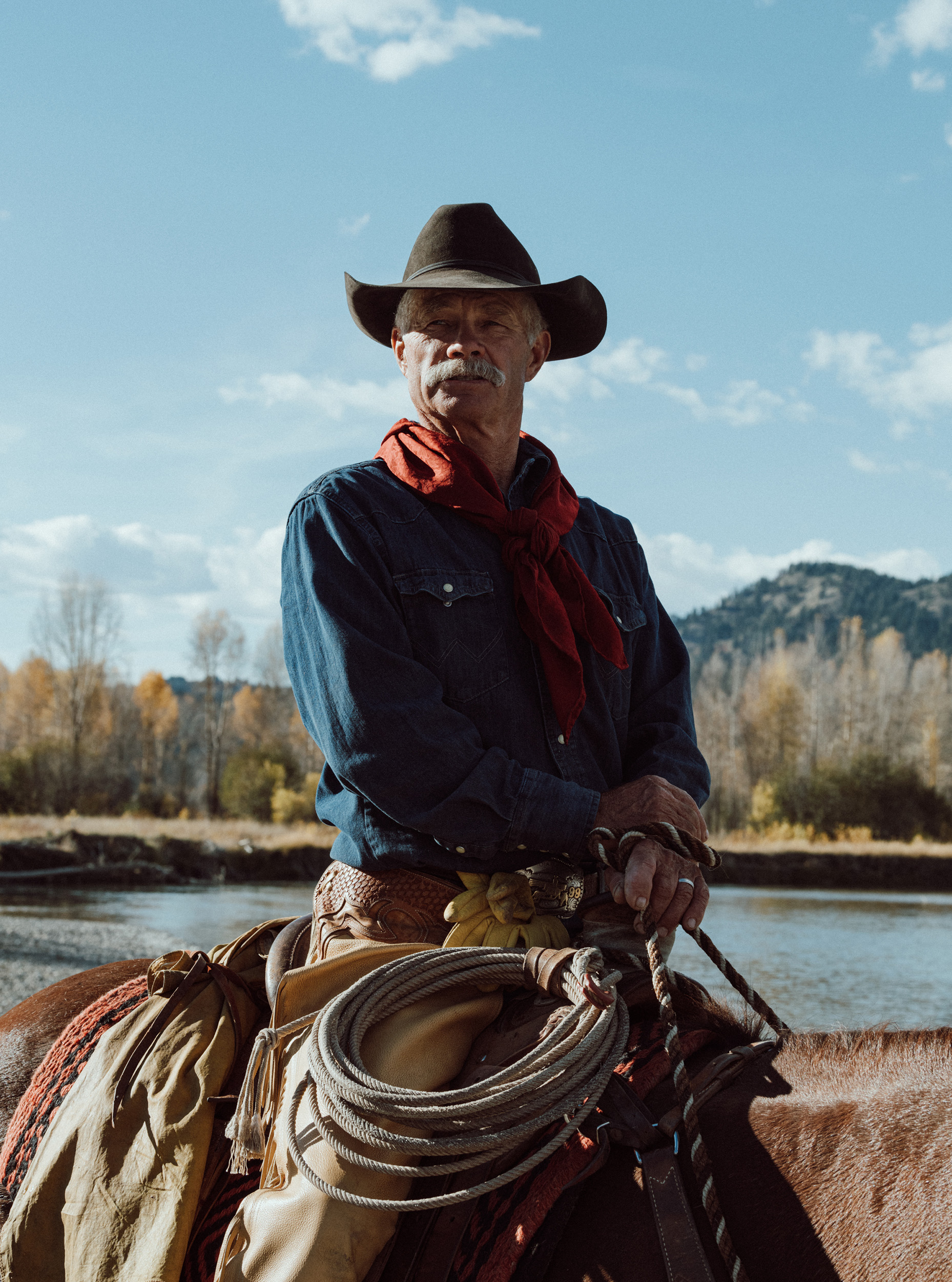 Portrait of a mature adult man on horseback wearing a cowboy hat and red scarf against a nature background of blue sky and water.