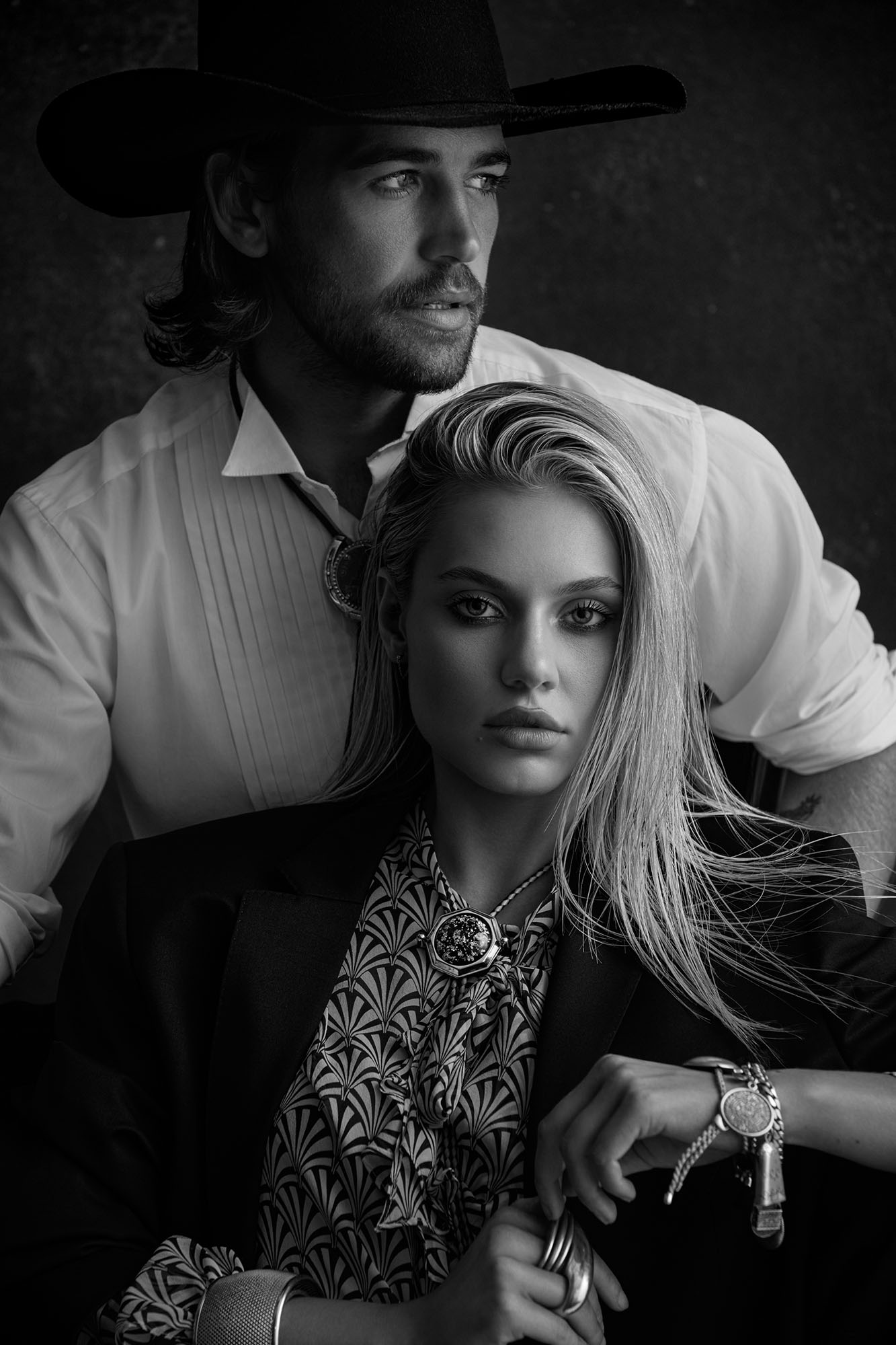 Black and white portrait of a woman in a patterned shirt and blazer with a man in a cowboy hat and white shirt behind her.
