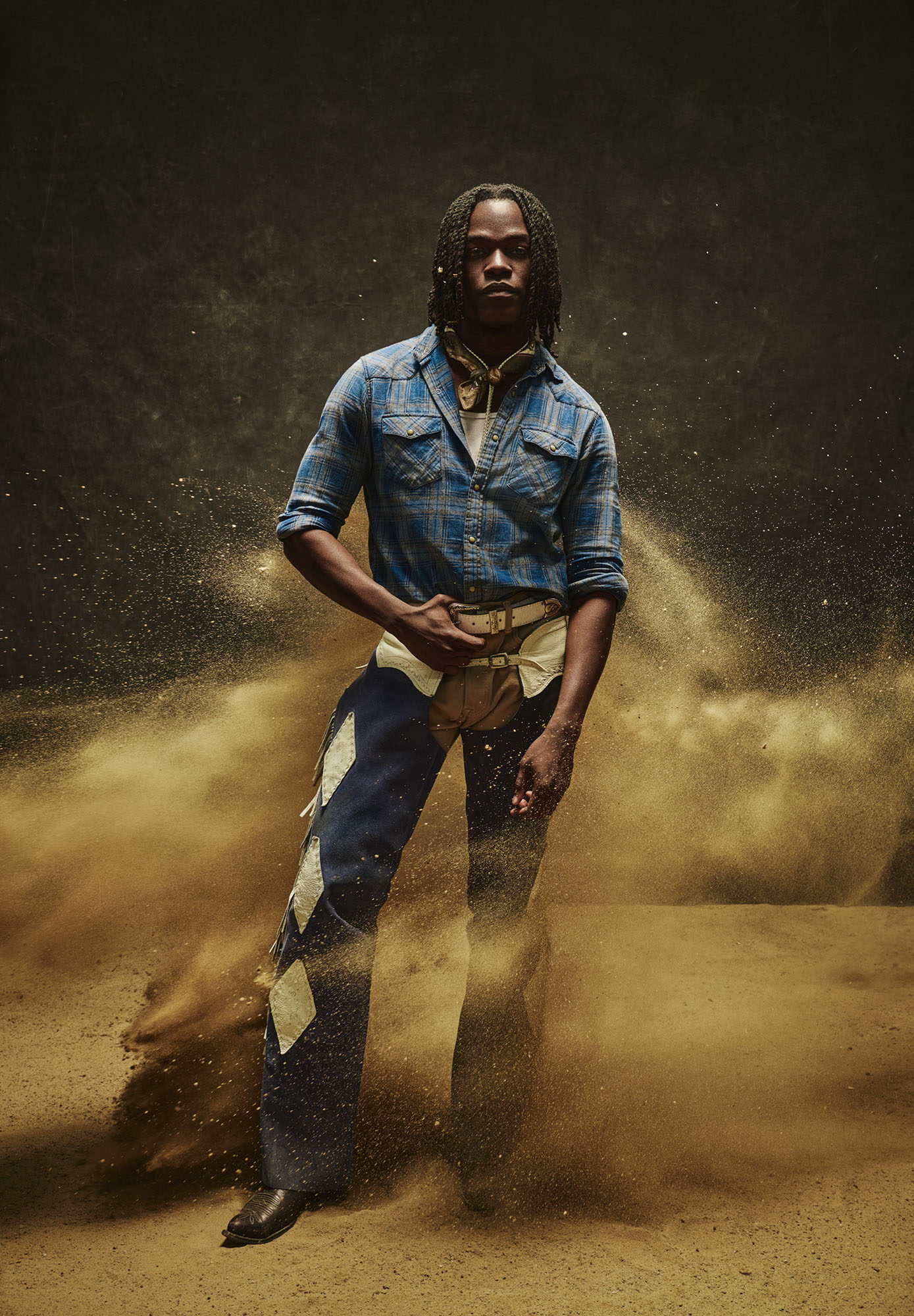 Portrait of a man with dreadlocks wearing a blue denim shirt, chaps, and boots, posing against a dark background with sand.