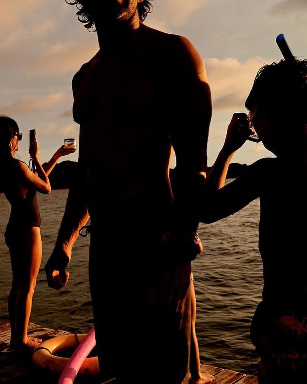 Three people silhouetted on a dock over water at sunset, one with snorkel gear, another taking photos, creating a warm, summery scene.