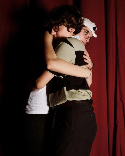Two people embrace in a dimly lit setting with a red curtain backdrop; one wears a white top and the other a striped polo shirt and baseball cap.