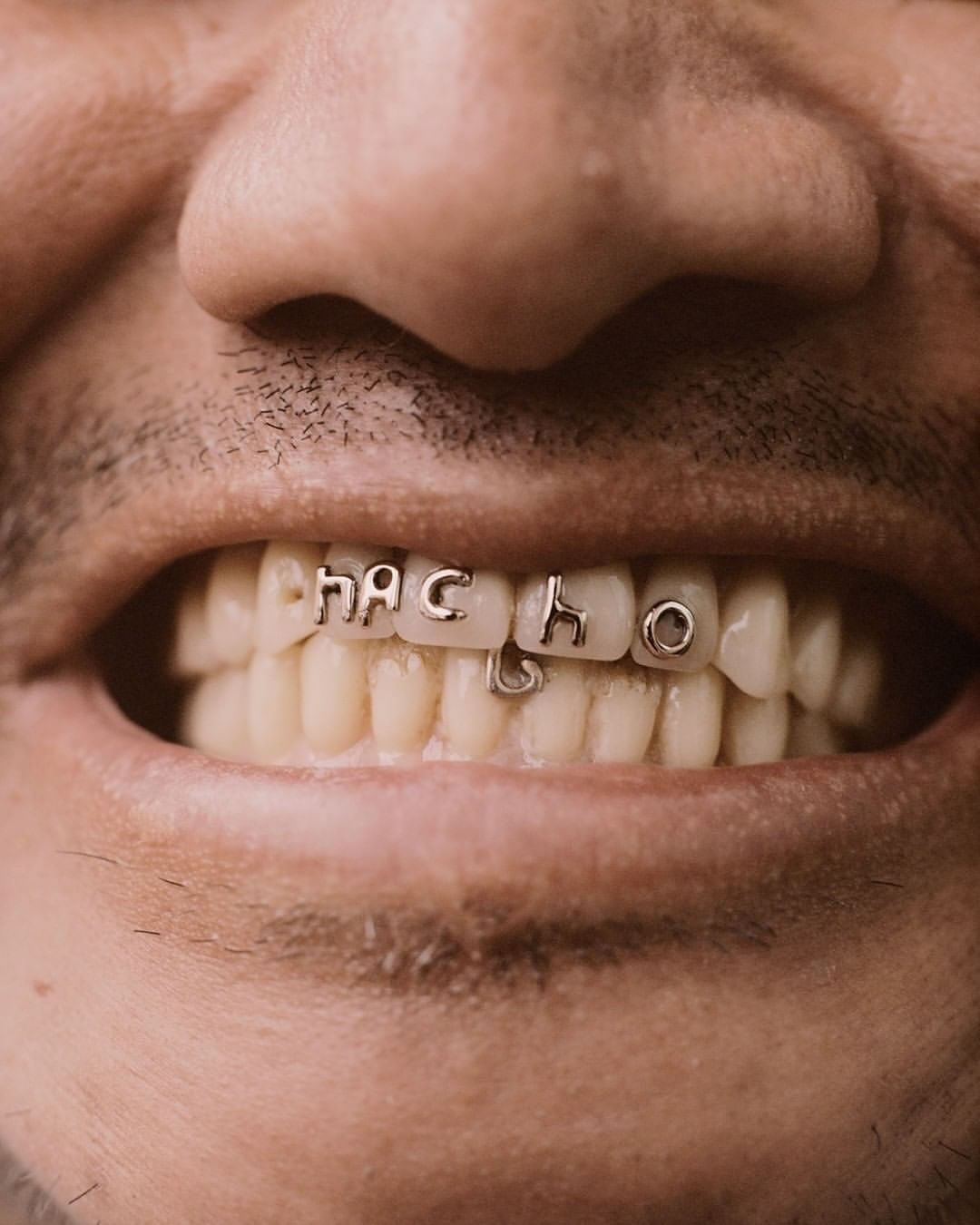Close-up of a man's mouth showing white teeth adorned with silver letters spelling 'NACHO' in a grill-like fashion.