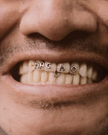 Close-up of a man's mouth showing white teeth adorned with silver letters spelling 'NACHO' in a grill-like fashion.