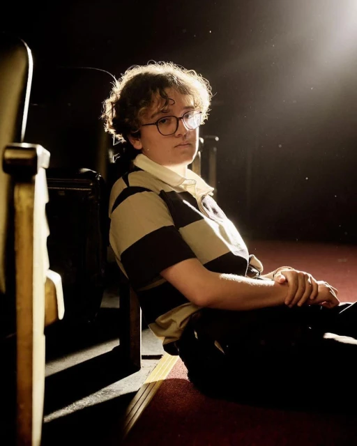 Portrait of a person with glasses sitting near theatre seating in a dimly lit space, illuminated by a spotlight.