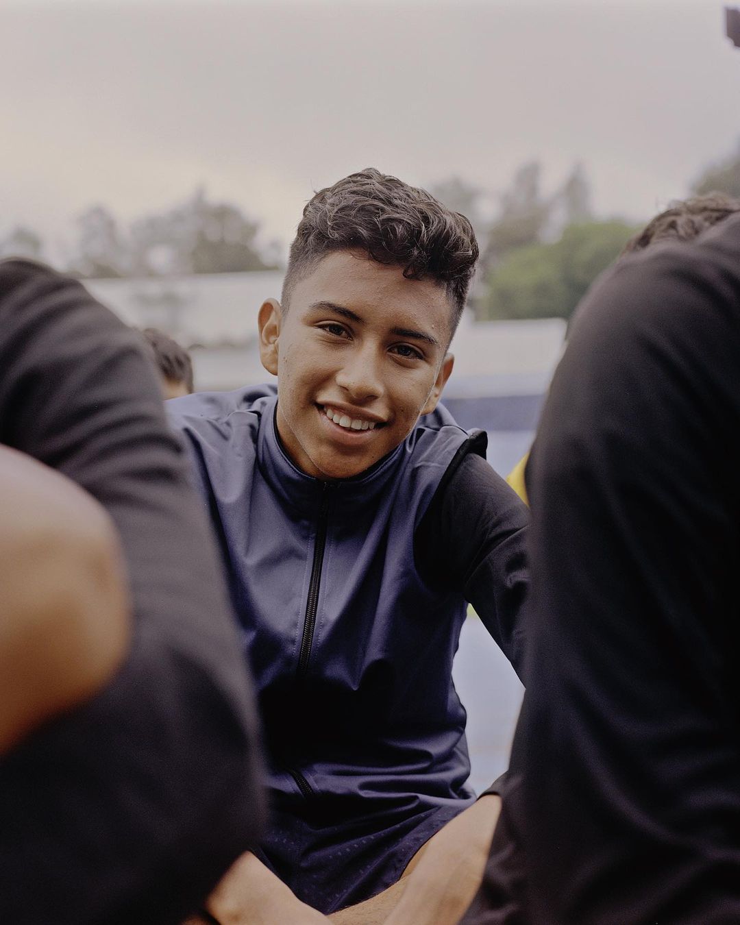 Smiling young man in athletic wear surrounded by teammates outdoors with a cloudy sky backdrop, showcasing youthful energy and camaraderie.