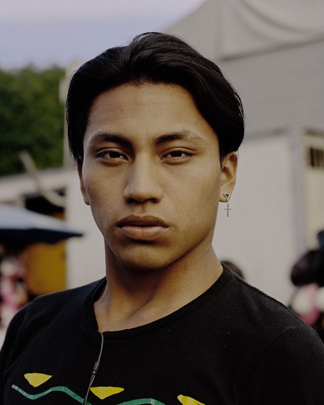 Portrait of an adult male with dark hair, wearing a black T-shirt with colorful geometric pattern and silver cross earring, medium shot.