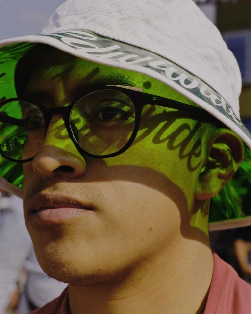 Close-up of a young adult with glasses wearing a bucket hat, face partially shadowed by the hat's brim, outdoor setting, natural light.