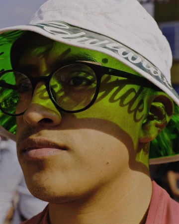 Close-up of a young adult with glasses wearing a bucket hat, face partially shadowed by the hat's brim, outdoor setting, natural light.
