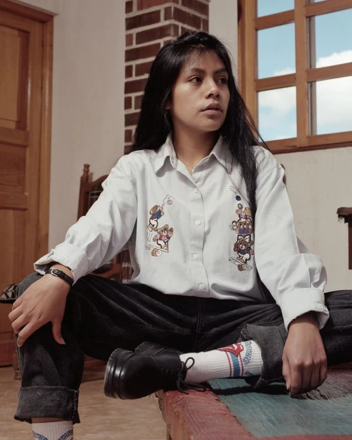 Young woman in a button-down shirt with cartoon bears sits on a bed near a window. She has long dark hair and is looking to the side.