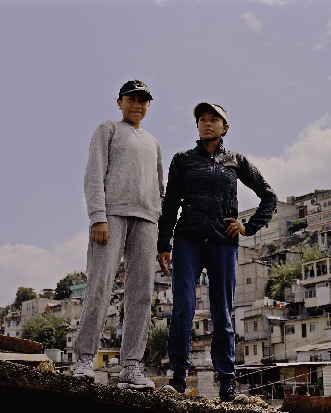Two young individuals stand together on a rooftop in sportswear, with an urban cityscape and a blue sky as their backdrop, casual style.
