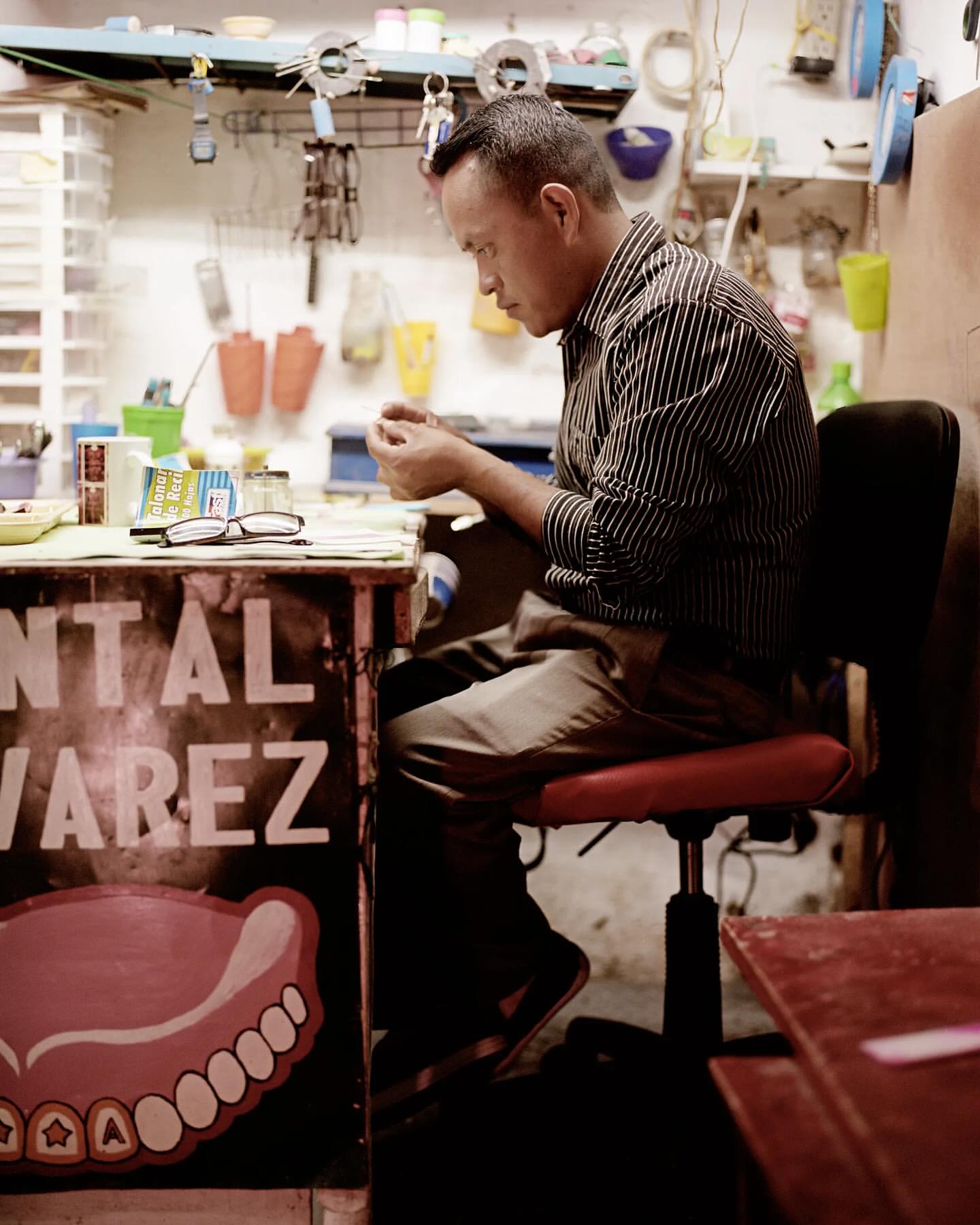 An adult male works at a desk in a small room. The subject is wearing a striped shirt and sitting on a red chair.