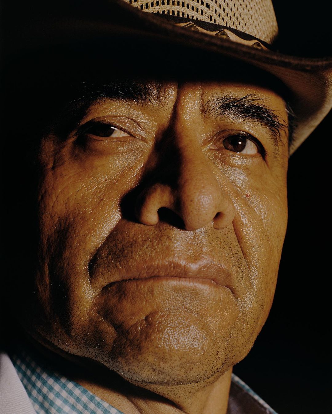 Close-up portrait of an adult man with a serious expression, wearing a cowboy hat, set against a dark background. 