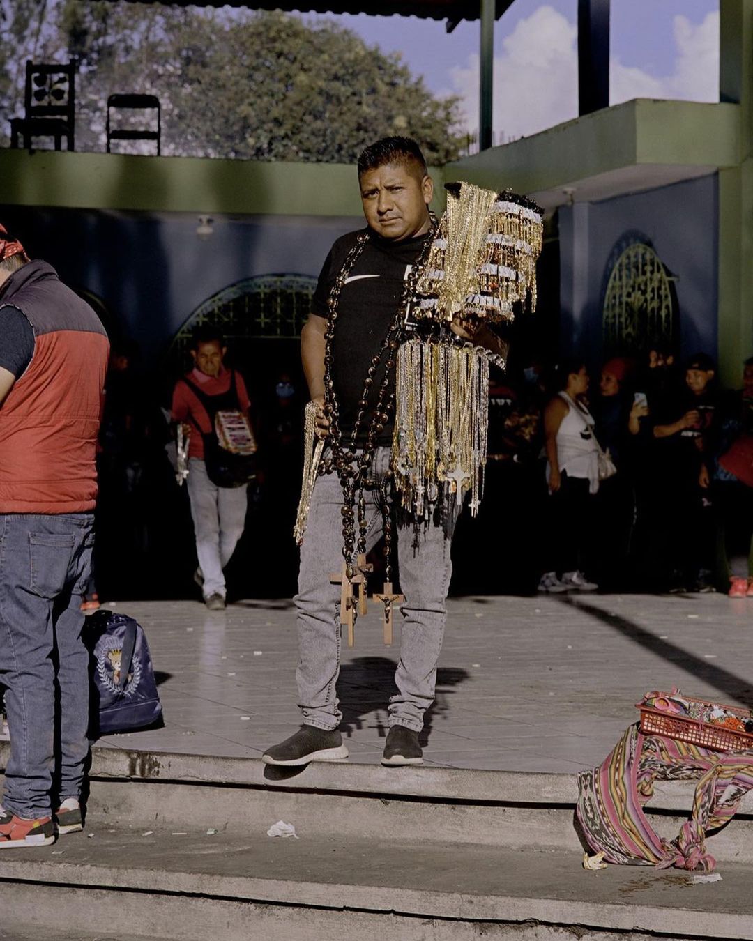 Adult male street vendor stands on steps with necklaces and rosaries in outdoor market setting with people in background during daytime.
