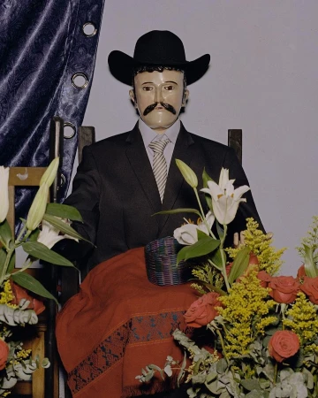 Mannequin dressed in a black suit and cowboy hat seated among floral arrangements including white lilies and red roses.