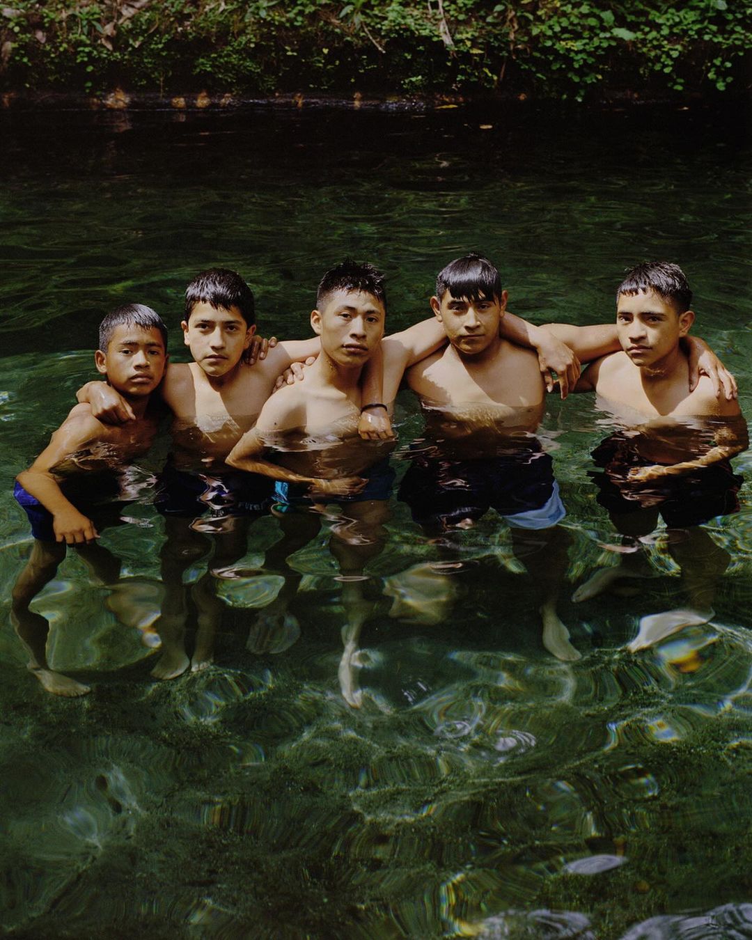 Five young Hispanic men stand in waist-deep, clear water surrounded by lush green vegetation in a natural setting.