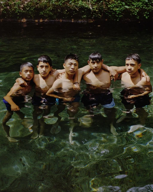 Five young Hispanic men stand in waist-deep, clear water surrounded by lush green vegetation in a natural setting.
