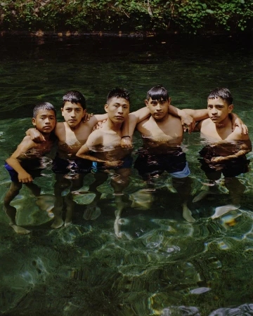Five young Hispanic men stand in waist-deep, clear water surrounded by lush green vegetation in a natural setting.