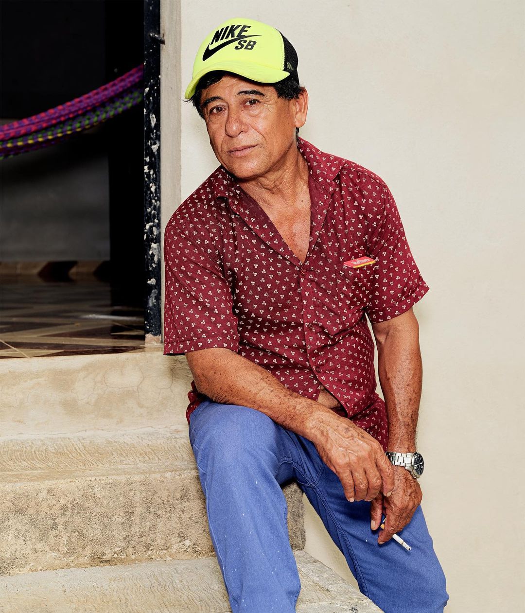 Older man in a patterned shirt and baseball cap sits on steps, holding a cigarette, captured in a medium shot with natural lighting.