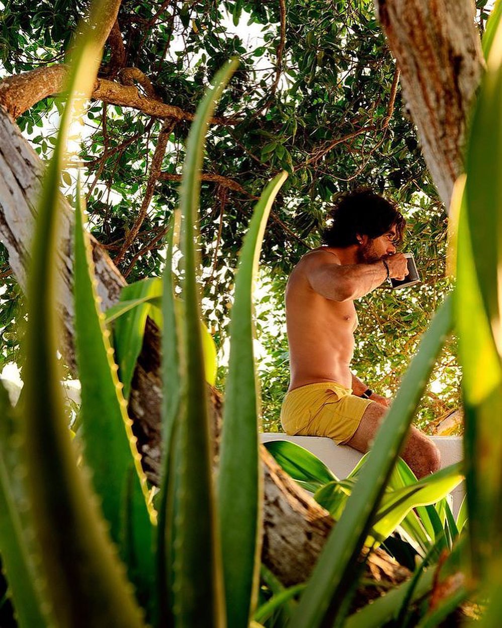 Adult man with dark hair and tan skin, shirtless in yellow shorts, sitting outdoors drinking from a black mug amongst green foliage.