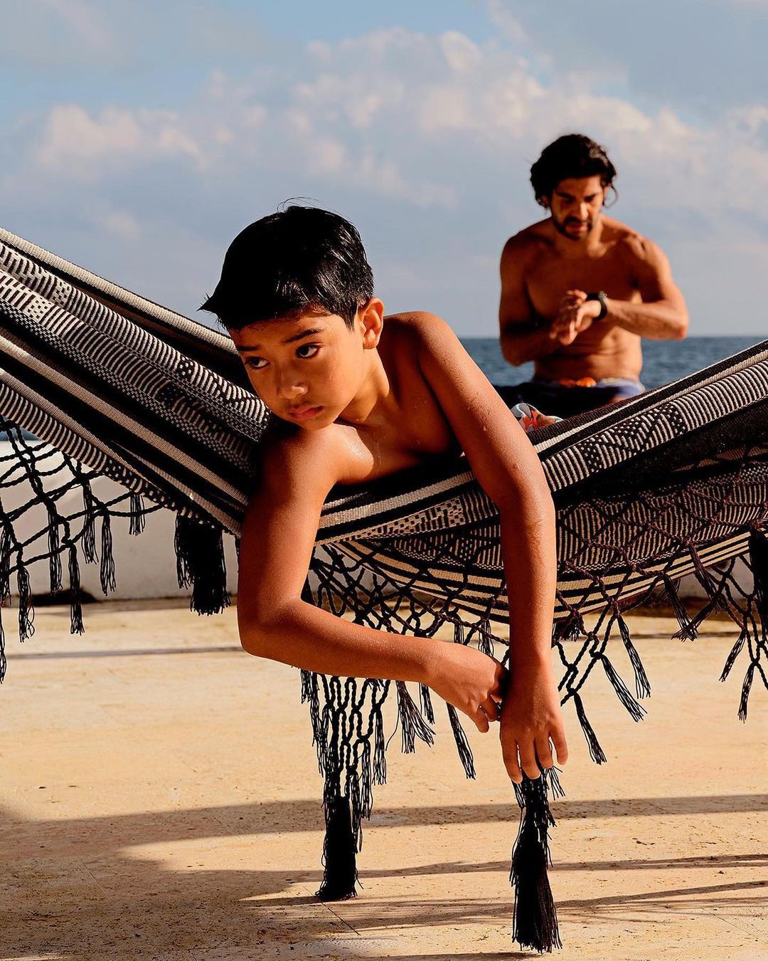 Child rests in a striped hammock outdoors with an adult male in the background against a beach and sky backdrop.