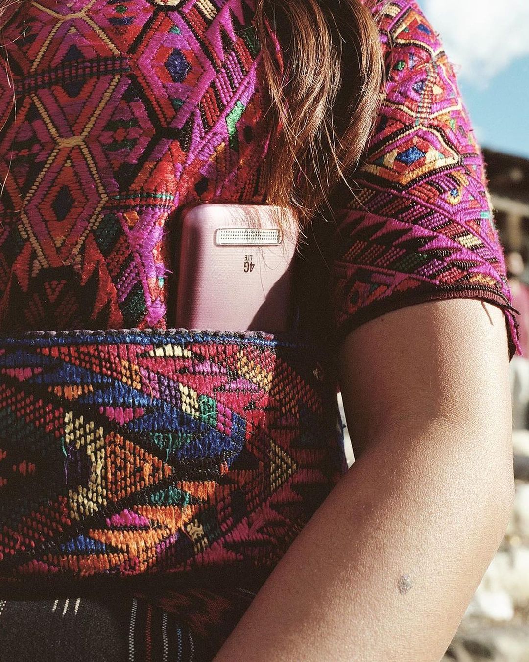 Close up of a woman with long brown hair wearing a patterned top and carrying a cell phone in a matching patterned bag.