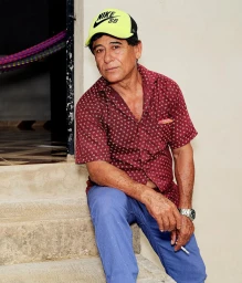 Man wearing a red patterned shirt, blue trousers, and lime green Nike SB cap sitting on concrete steps holding a cigarette