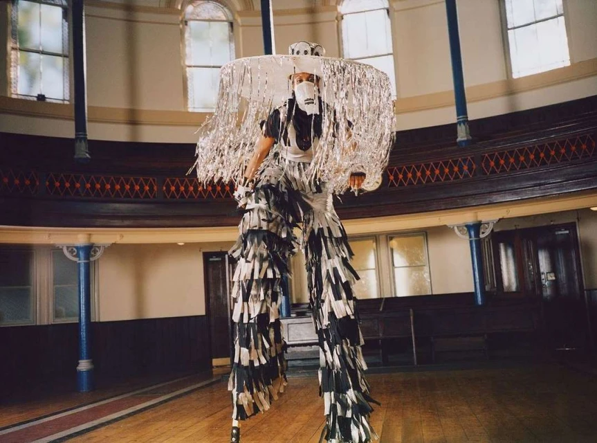 Performer on stilts wearing a silver fringe hat and mask in a performance space with wood floors and large windows.