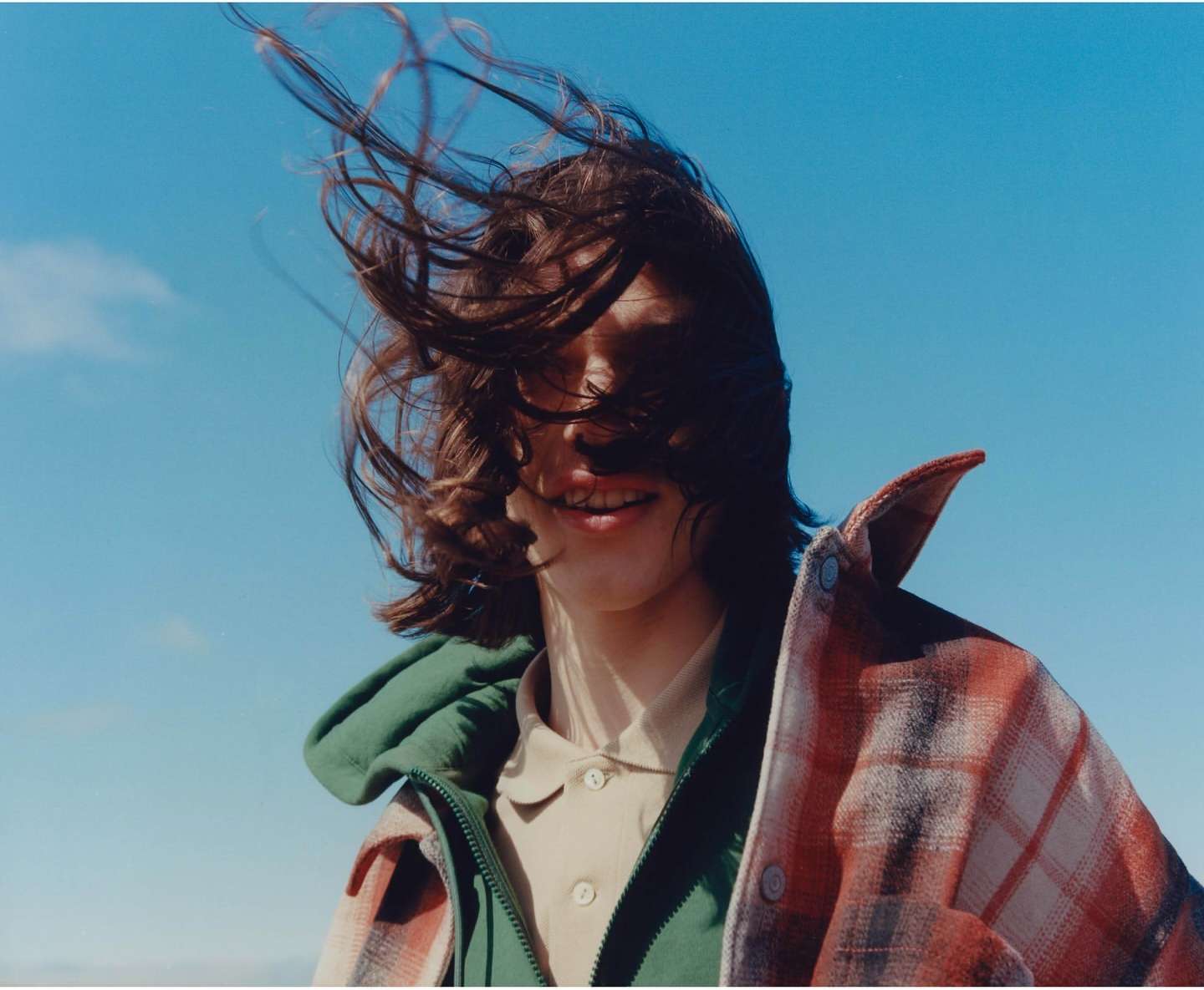 An adult with wind-blown hair wears a green hooded jacket and a plaid shirt against a clear blue sky in an eye-level outdoor shot.