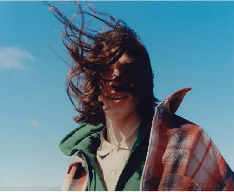 An adult with wind-blown hair wears a green hooded jacket and a plaid shirt against a clear blue sky in an eye-level outdoor shot.