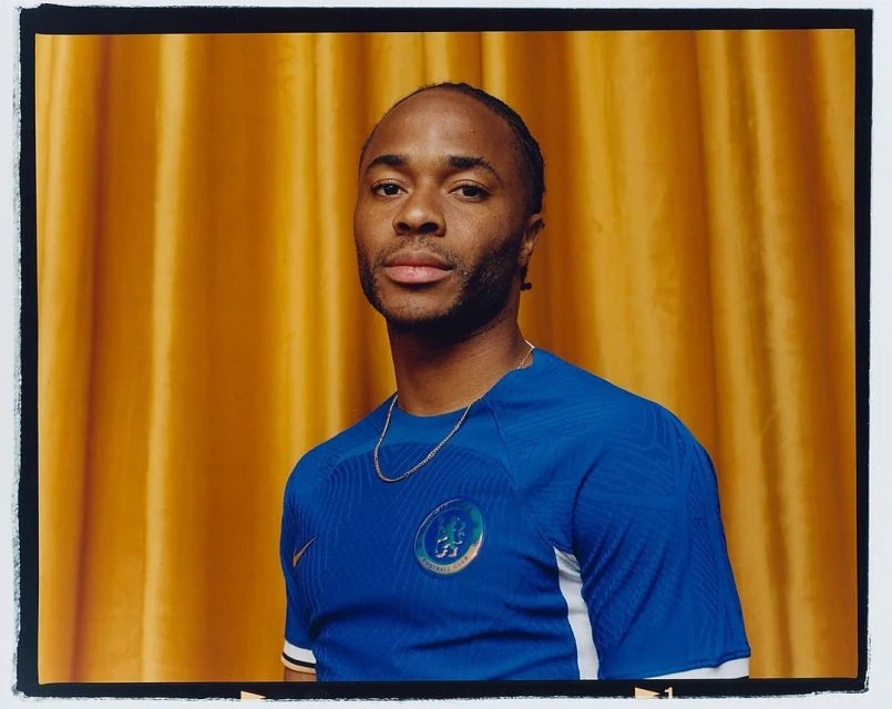 Portrait of a man with cornrows in a blue soccer jersey with a gold chain against a golden yellow curtain backdrop.