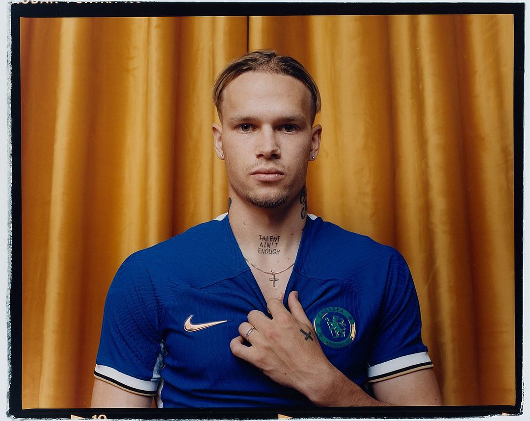 Blond man in blue soccer jersey stands against gold curtain, showcasing tattoos, cross necklace, ring, and a confident, neutral expression.