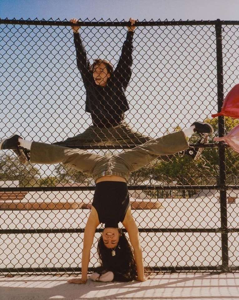Two people performing acrobatics on a fence outdoors on a sunny day. One woman is upside down with the other person balanced above her.