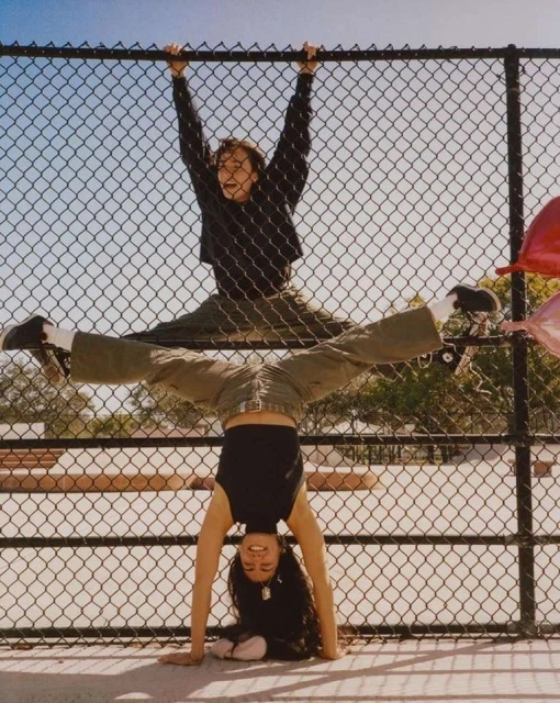 Two people performing acrobatics on a fence outdoors on a sunny day. One woman is upside down with the other person balanced above her.