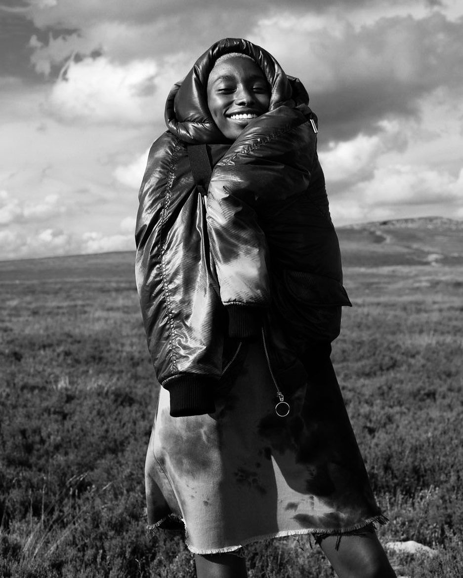 Black and white fashion shot of a smiling woman in oversized puffer coat standing in a grassy, rural landscape under a cloudy sky.