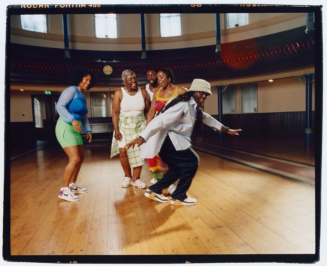 Group of five people dancing indoors on a wooden floor, wearing casual clothing and sneakers, in a well-lit space with large windows.