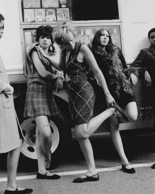 Five women in 1970s-style fashion are gathered outdoors beside a truck, showcasing vintage clothing and accessories in a monochrome editorial style.