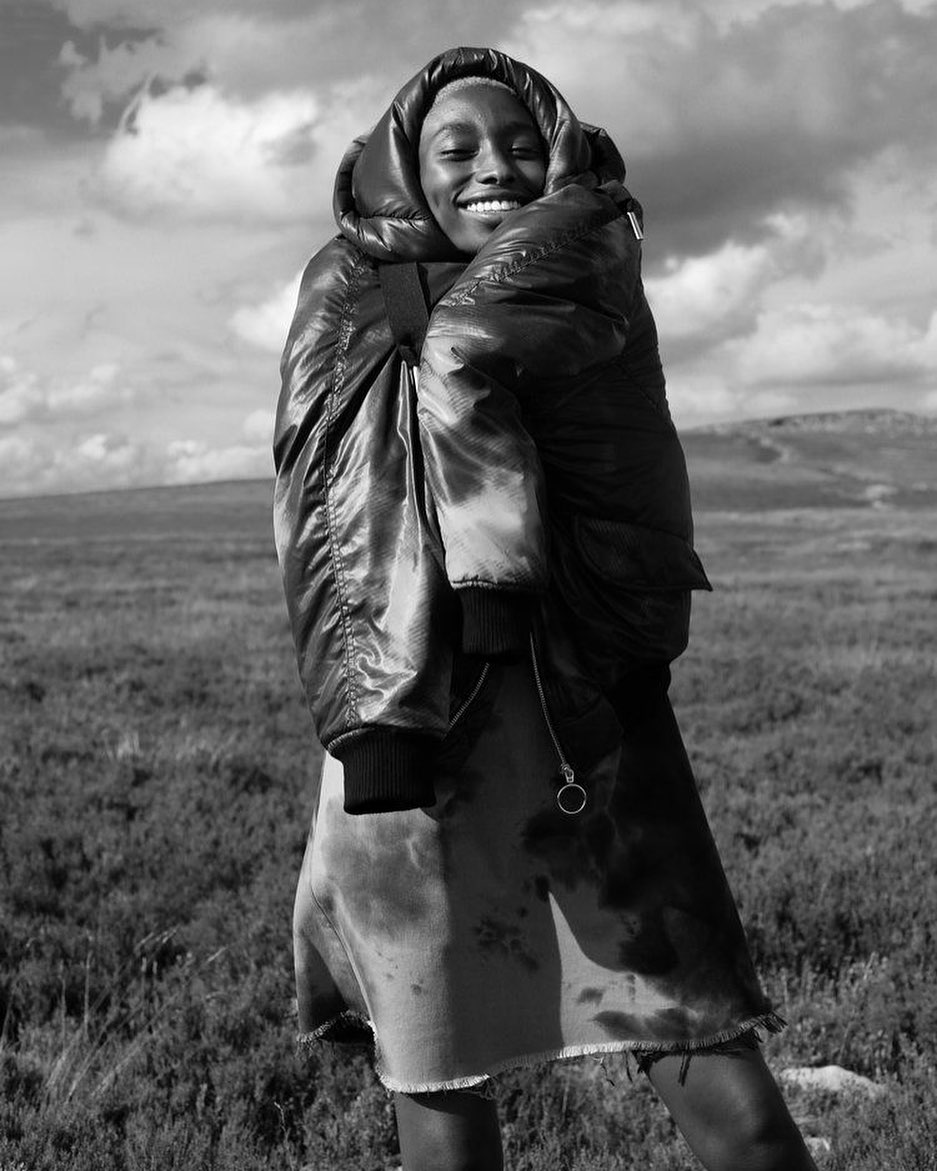Monochrome portrait of a smiling African woman outdoors wearing a puffy jacket and skirt, against a field and cloudy sky.