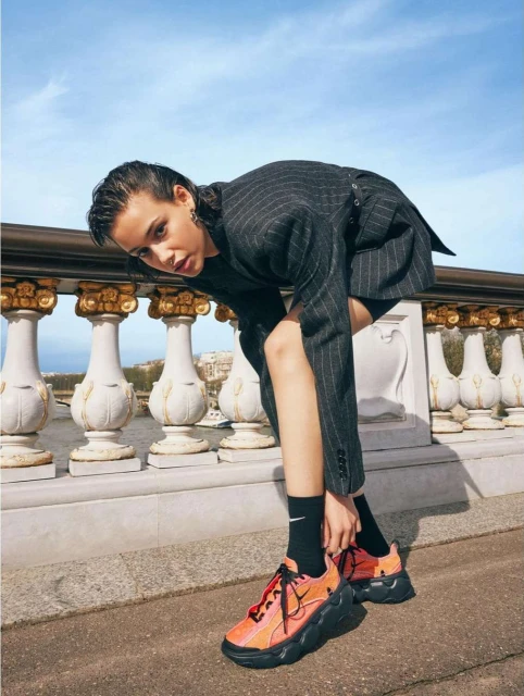 Woman in pinstripe blazer tying orange and black sneakers on a stone bridge with decorative balustrade, with a blue sky background.