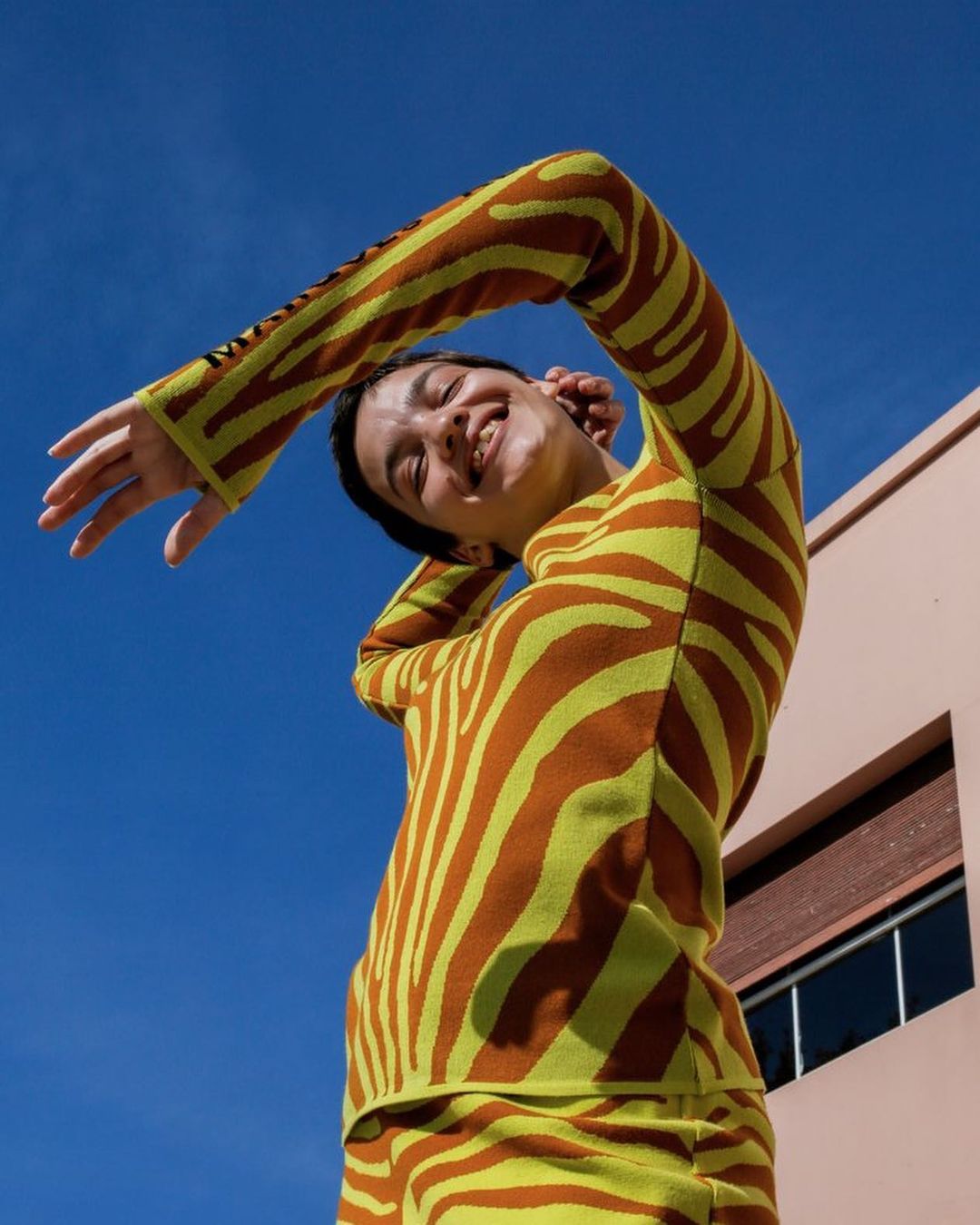 Smiling woman in a zebra-patterned outfit poses against a blue sky and the corner of a peach-colored building on a sunny day.
