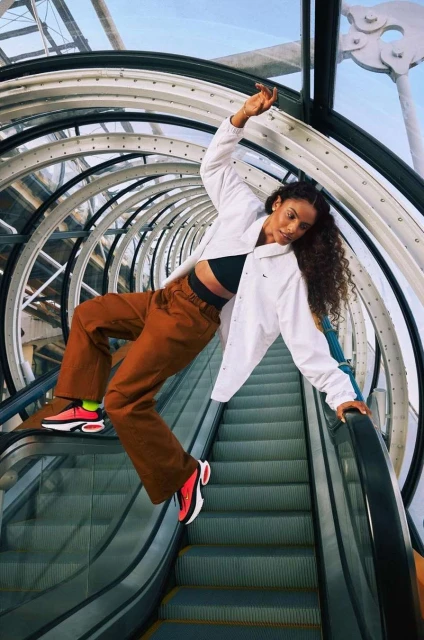 Athletic woman in a white jacket and brown pants poses on an escalator in a circular tunnel, showcasing red and black sneakers.