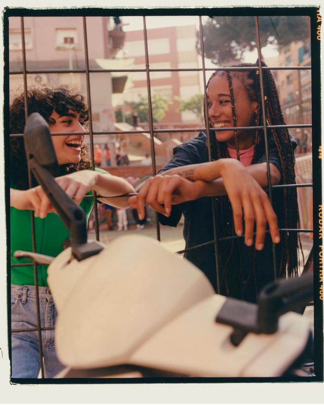 Two women laugh together, leaning on a metal fence with an out-of-focus scooter in front. Outdoor scene with urban buildings in the background.