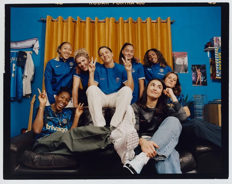 Group portrait of eight smiling adult women in blue sports jerseys, posing on a couch in front of a blue wall and yellow curtain.