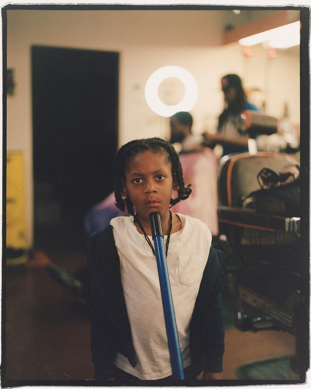 A young Black child stands in a barbershop, holding a blue mop, with other people and barber chairs blurred in the background.