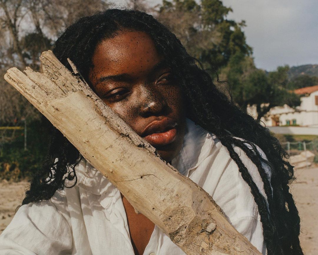 Portrait of a Black woman with long braids, embracing a piece of driftwood on a sandy beach with blurred background of trees and a building.