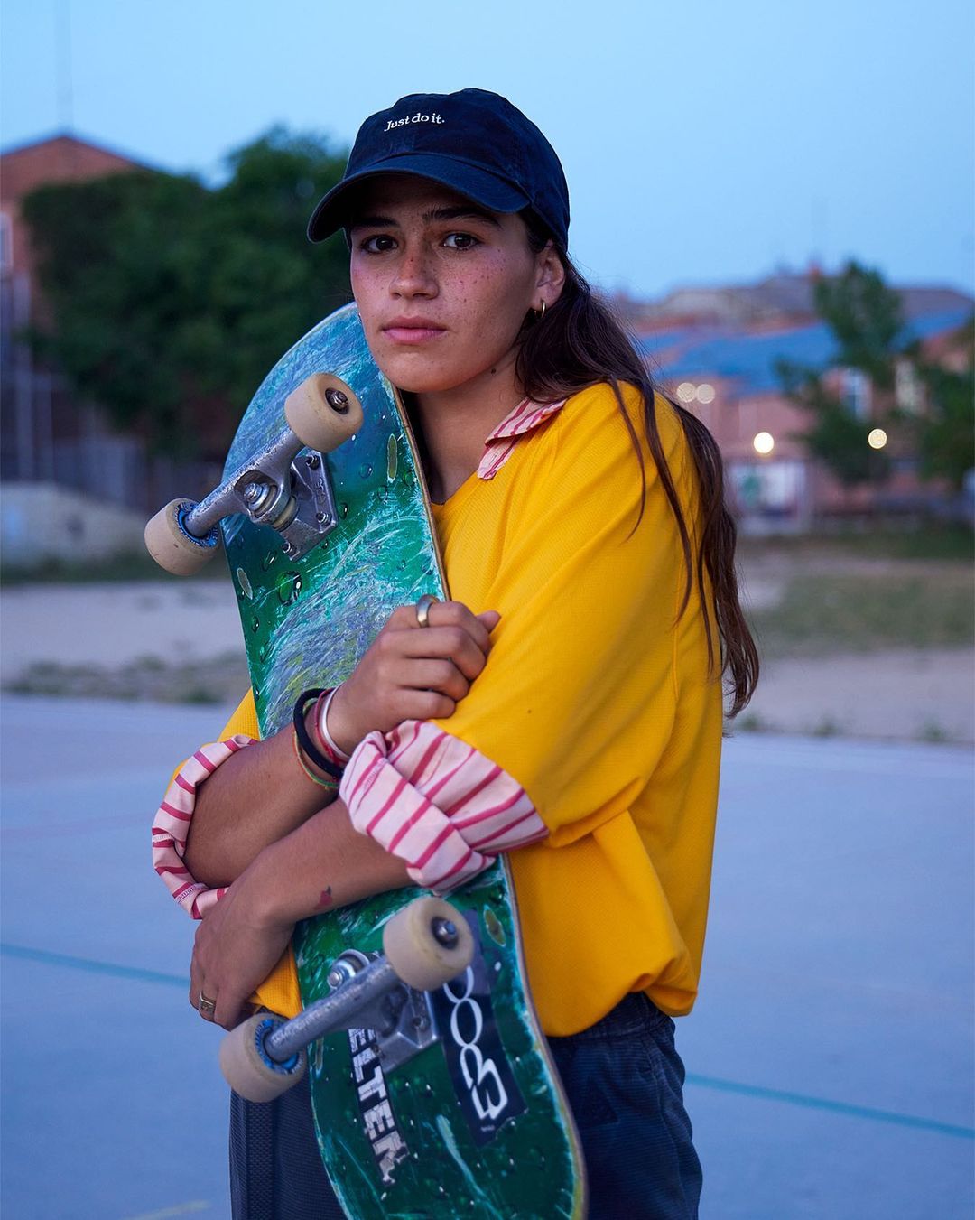 Young woman with a skateboard poses outdoors at dusk, wearing a yellow shirt and cap. The image evokes urban lifestyle and sport.