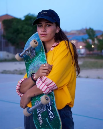 Young woman with a skateboard poses outdoors at dusk, wearing a yellow shirt and cap. The image evokes urban lifestyle and sport.