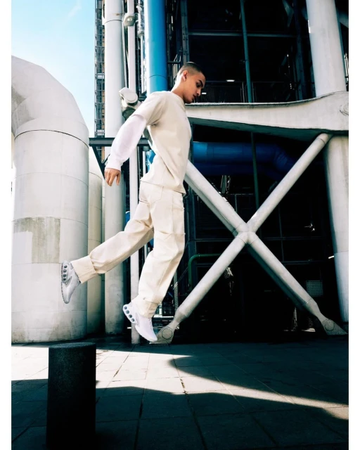 A person in a light-colored outfit is captured mid-air in a jump, set against an industrial urban backdrop of pipes and structural beams.