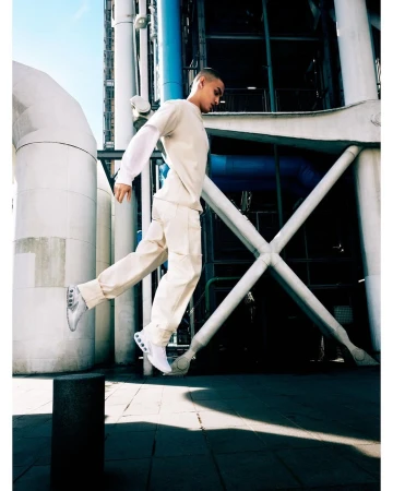 A person in a light-colored outfit is captured mid-air in a jump, set against an industrial urban backdrop of pipes and structural beams.