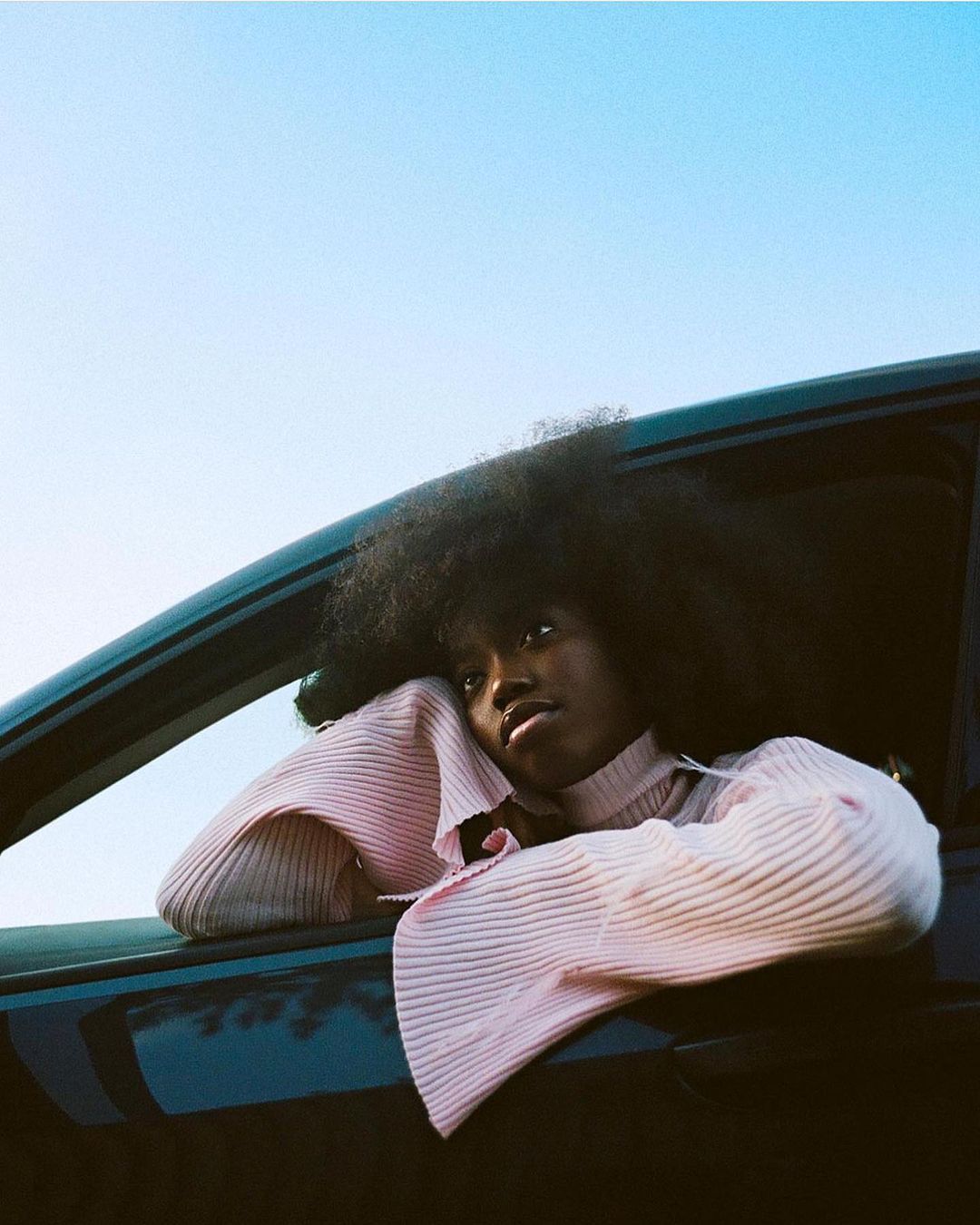 A young woman with afro rests her arms on a car window frame against a light blue sky, creating a fashion-forward and relaxed visual.